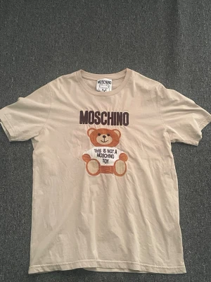 Beige Moschino t-shirt med björntryck - Säljer en beige t-shirt från Moschino med ett stort tryck av en nallebjörn och texten 'This is not a Moschino toy' på framsidan. T-shirten har rund hals och korta ärmar. Perfekt för dig som gillar statement-plagg och coola prints. Passar även XL 