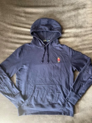Polo Ralph Lauren Hoodie Navy – Polo Bear, Storlek M - Snygg mörkblå hoodie med huva och dragsnören. På bröstet finns en liten broderad figur i färg som ger en unik touch. Hoodien har en klassisk känguruficka framtill och är tillverkad i mjukt material, perfekt för en avslappnad stil.
