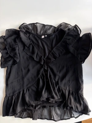 Svart volangblus med kort ärm - Snygg svart blus i transparent, lätt material med volangdetaljer längs ärmar, halsringning och nederkant. Blusen har korta ärmar och knäppning framtill. 