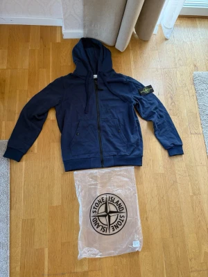 Mörkblå hoodie från Stone Island - Snygg mörkblå hoodie från Stone Island med dragkedja framtill och klassisk logopatch på vänster ärm. Hoodien har två fickor med knappstängning och ribbade muddar vid ärmslut och nederkant. Perfekt för en avslappnad streetwear-look.