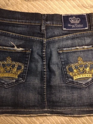Victoria Beckham kjol - Säljer en mörkblå jeanskjol med rå kant och snygga guldbroderade kronor på bakfickorna. Kjolen har klassisk femficksdesign och tydliga sömmar. Perfekt för dig som gillar detaljer och streetstyle.