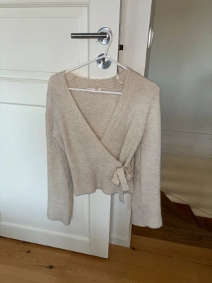 Beige omlottstickad tröja - Mysig beige stickad omlott-tröja med knytband i sidan och långa ärmar. Tröjan har en djup V-ringning och är gjord i ett mjukt, fluffigt material som känns skönt mot huden. Perfekt för lager på lager och en avslappnad men stilren look. snygg, kom gärna med bud och frågor ❤️