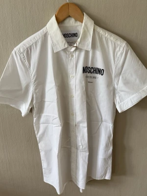 Vit kortärmad skjorta från Moschino - Snygg vit skjorta från Moschino med korta ärmar och klassisk krage. Skjortan har tryckt logga på bröstet och knäppning framtill. Perfekt för en clean och stilren look.0086