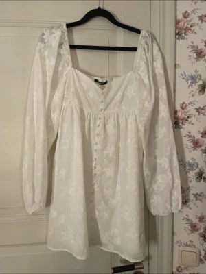 Vit blommig studentklänning - Söt vit studentklänning med blommigt mönster, långa puffärmar och knappar framtill. Klänningen har fyrkantig ringning och smock i ryggen för extra komfort. Perfekt för sommarens alla tillfällen! Jättejättefin klänning som jag tänkte använda på studenten men jag valde en annan istället så denna är i helt nyskick! Passar S, köpte den först i S men den satt lite mer som en M.