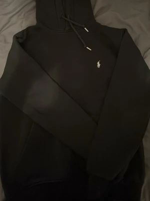 Svart hoodie från Polo Ralph Lauren - Snygg svart hoodie från Polo Ralph Lauren med klassisk vit broderad logga på bröstet. Hoodien har dragsko i huvan och en stor magficka. Tillverkad i mjukt material som känns skönt mot huden. Perfekt för en avslappnad och stilren look.