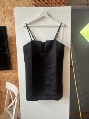 Svart kort klänning - Snygg svart kort klänning med smala axelband och rak passform. Klänningen har en minimalistisk design och är perfekt för dig som gillar stilrena plagg. Tillverkad i ett slätt material som ger en elegant look.