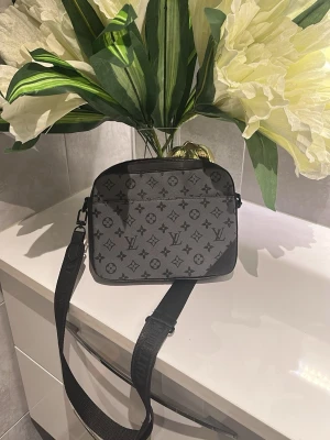 Grå axelväska med monogram sällsynt modell  - Snygg grå axelväska med svart monogrammönster och justerbar bred axelrem. Väskan har en rektangulär form och dragkedja upptill. Perfekt för dig som vill ha en stilren och trendig accessoar till vardagslooken.