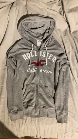 Grå zip hoodie från Hollister - Snygg grå hoodie från Hollister med dragkedja, vit text och röd logga på bröstet. Mjuk bomullsmix, två fickor fram och justerbar huva med vita snören. Perfekt för chill dagar eller när du vill ha en casual look.