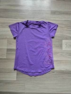 Lila sport-tshirt med tryck - Lila sport-tshirt i syntetmaterial med korta ärmar och rund hals. Framsidan har ett rosa tryck längs ena sidan och nederkanten är rundad både fram och bak. Perfekt för träning och aktivitet.
