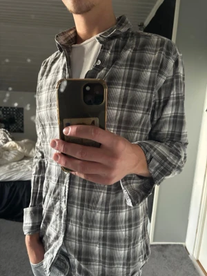 Rutig skjorta - Säljer en grå rutig skjorta med klassisk krage och knäppning framtill. Skjortan har lång ärm och normal passform, perfekt att bära över en t-shirt för en avslappnad look.