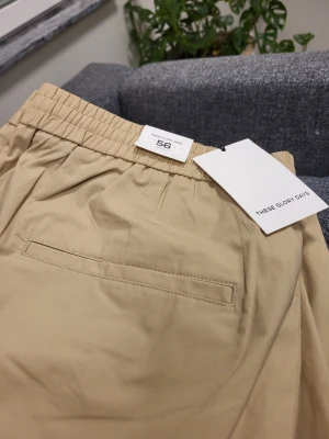 Beige chinos från These Glory Days - Snygga beige chinos från These Glory Days i storlek 56. Byxorna har en rak passform, elastisk midja och klassiska fickor både fram och bak. Tillverkade i ett mjukt och lätt material som ger en clean och stilren look.