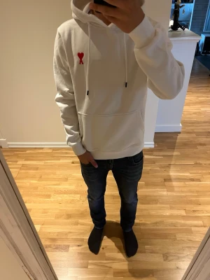 Vit hoodie med rött hjärta - Säljer en stilren vit hoodie med huva och dragsnören. På bröstet finns ett litet rött hjärta med bokstaven A. Hoodien har en stor magficka och ribbade muddar vid ärmar och nederkant. Perfekt för en avslappnad och trendig look.