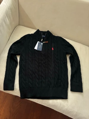 Svart kabelstickad tröja Polo Ralph Lauren - Kabelstickad svart tröja från Polo Ralph Lauren med halv dragkedja och ribbade muddar. Tröjan har en liten röd logga broderad på bröstet och är tillverkad i ett mjukt material. Perfekt för dig som gillar klassisk och stilren look.