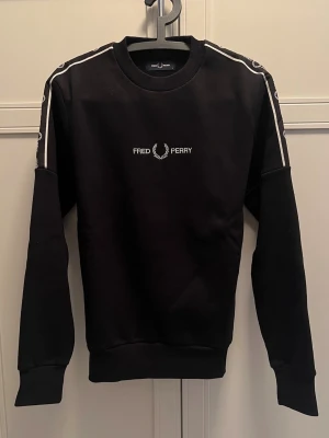Svart sweatshirt från Fred Perry - Svart sweatshirt från Fred Perry med vita detaljer på ärmarna och logga broderad på bröstet. Klassisk rund halsringning och ribbade muddar vid ärmslut och nederkant. Perfekt för en stilren look.