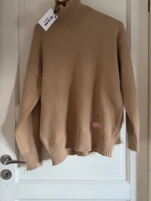 MEET ME THERE polo tröja  - Beige stickad tröja från MEET ME THERE med hög krage och ribbade muddar. Tröjan har en söt liten rosa körsbärsdetalj broderad nedtill. Oversized passform och mjukt material som känns skönt mot huden.