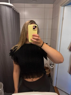 Svart croppad topp med volang - Säljer en svart croppad topp med kabelstickat mönster och volangdetaljer i mesh vid ärmar och nederkant. Toppen har korta ärmar och en rund halsringning. Perfekt för dig som vill ha en trendig och unik look.