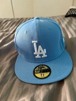 Ljusblå LA 59FIFTY keps - Snygg ljusblå 59FIFTY keps från New Era med LA-logga broderad i vitt framtill. Klassisk flat brim och strukturerad passform. Kepsen har även New Era-loggan broderad på sidan. Perfekt för dig som gillar streetwear och sportig stil.