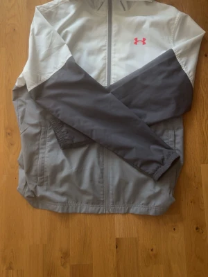  Under Armour jacka - Snygg vindjacka från Under Armour i grått och vitt med rosa logga på bröstet. Jackan har hel dragkedja framtill, huva och långa ärmar. Tillverkad i lätt syntetmaterial som andas, perfekt för blåsiga dagar.