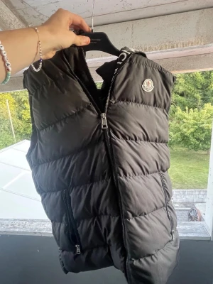 Svart Moncler väst - Snygg svart dunväst från Moncler med dragkedja framtill, två fickor med dragkedja och Moncler-logga på bröstet. Insidan har ett band med Moncler-tryck. Perfekt för lager-på-lager och streetstyle.