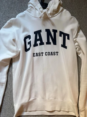 Vit hoodie från GANT med tryck - Snygg vit hoodie från GANT med svart text 'GANT EAST COAST' på bröstet. Klassisk huva med dragsko och mjukt material som känns skönt mot huden. Perfekt för dig som gillar en clean och sportig stil.