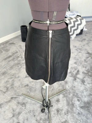 Svart skinnkjol från Gina Tricot - Snygg svart kort skinnkjol från Gina Tricot med en stor silverfärgad dragkedja framtill. Kjolen har en clean look och passar perfekt till en trendig outfit. Materialet är mjukt och följsamt syntetiskt skinn.
