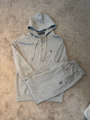 Grey Polo Ralph Lauren Tracksuit - This is a matching grey tracksuit set from Polo Ralph Lauren. It includes a zip-up hoodie and jogger pants, both featuring the iconic Polo Ralph Lauren embroidered logo in black. The material appears to be a soft, comfortable knit, suitable for casual or loungewear. (Pants in M)