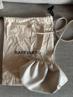 Beige axelväska med dragsko - Snygg beige axelväska från RAFFINATO, som ni ser på bilderna är den lite sliten och har små små blåa prickar inuti väskan, min inget man tänker på. Man kan även använda denna som clutch väska. Man får tygpåsen till.