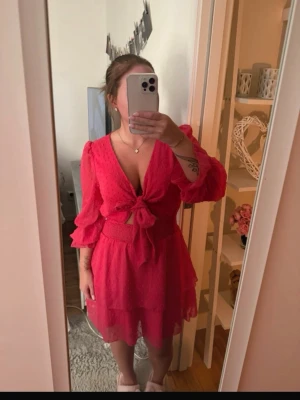 Rosa kort klänning med knyt framtill - Supersöt rosa kort klänning med v-ringning, knyt framtill och volangdetaljer. Klänningen har trekvartsärm och är tillverkad i ett lätt, prickigt tyg som ger en luftig känsla. Perfekt för sommarens alla tillfällen!
