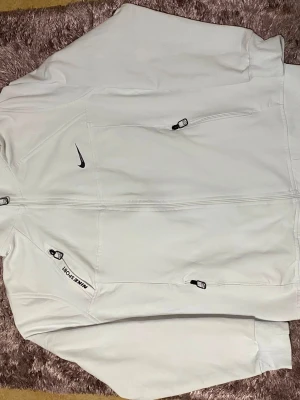 Vit hoodie från Nike Sport - Snygg vit hoodie från Nike Sport med dragkedja framtill och flera praktiska fickor med dragkedja. Diskret Nike-logga på bröstet och texten NIKE SPORT på ena fickan. Tillverkad i mjukt material och har ribbade muddar vid ärmslut och nederkant.