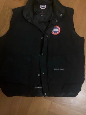 Svart dunväst från Canada Goose - Säljer en svart dunväst från Canada Goose med klassisk logga på bröstet och två stora fickor framtill. Västen har hög krage och stängs med tryckknappar och har en dragkedja. Perfekt för lite kyliga dagar nu till hösten och vintern och riktigt snygg till lager-på-lager-stil. Pris kan diskuteras vid snabb affär