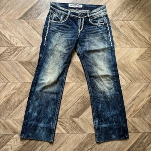 Y2K jeans från grasshopper - Säljer ett par blå bootcut jeans med tydliga slitningar och ljusa tvättade partier. Jeansen har dekorativa sömmar och detaljer vid fickorna samt raka ben. Perfekta för en avslappnad och trendig look.