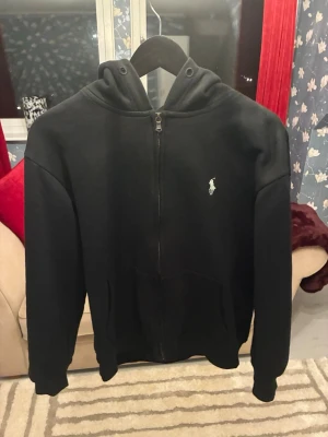 Svart hoodie från Polo Ralph Lauren - Svart hoodie från Polo Ralph Lauren med vit broderad logga på bröstet. Tröjan har dragkedja framtill, huva och är tillverkad i mjuk bomull. Står S men passar mycket bra till XS.                              Måtten på tröjan är som följande: Framtill (dragkedjans längd) :54cm. Armlängd (Från armhålan) :45cm. Från Axelsömmen till andra axellsömmen: 50cm.