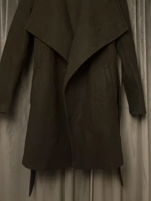 Svart kappa med knytband - Stilren svart kappa med bred krage och knytband i midjan. Kappan har långa ärmar och två sidofickor. Materialet ser ut att vara ullblandning, perfekt för kyligare dagar. Enkel och tidlös design som passar till många outfits.