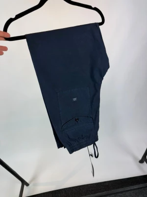 Mörkblå cargobyxor med dragsko - Snygga mörkblå cargobyxor med praktisk ficka på benet och dragsko vid benslut. Byxorna har en avslappnad passform och är perfekta för en streetwear-look. Tillverkade i ett slitstarkt material som passar till många olika stilar.