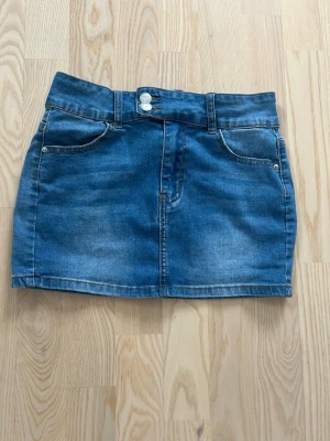 Blå jeanskjol med fickor - Snygg blå jeanskjol i klassisk modell med två framfickor och två bakfickor. Kjolen har bälteshällor och stängs med knapp och dragkedja framtill. Perfekt till vardags för en avslappnad och trendig look.