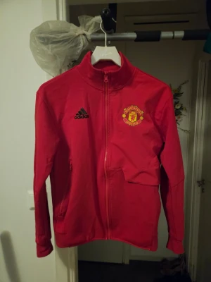 Röd Manchester United träningsjacka - Snygg röd träningsjacka från Adidas med Manchester Uniteds klubbmärke på bröstet. Jackan har hel dragkedja, hög krage och två sidofickor med dragkedja. Perfekt för fotbollsträning eller att bära till vardags.