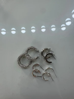 Earrings Collection - This image shows four pairs of hoop earrings arranged on a flat surface. The top left pair is encrusted with small stones, giving them a sparkly appearance. The other three pairs are plain metallic hoops of varying sizes and thicknesses.