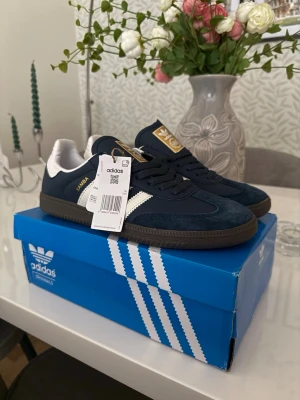 Adidas Samba OG marinblå sneakers - Snygga Adidas Samba OG sneakers i marinblått med vita detaljer och guldfärgade loggor. Skorna har klassisk låg profil, ovandel i mocka och läder samt gummisula. Perfekta för dig som gillar retrostil och streetwear.