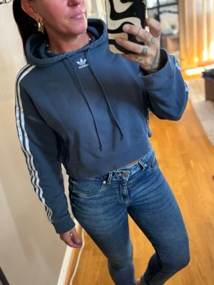Blå cropped hoodie från Adidas - Snygg blå cropped hoodie från Adidas med klassiska vita ränder längs ärmarna och Adidas-logga på bröstet. Hoodien har huva med dragsko och ribbade muddar vid ärmsluten. Perfekt för en sportig och avslappnad stil. Storlek 34 men jag har S och passar bra