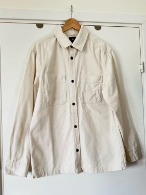 H&M - vit overshirt / skjorta i 100% bomull - storlek M - Stilren vit skjorta / overshirt från H&M i storlek M. Tidlös modell med klassisk krage, långa ärmar och krage och två bröstfickor. Knäpps med svarta knappar fram. Stabil grovvävd denim / jeans i 100% bomull, så bra naturmaterial som både ger stadga och andas. Lika snygg som skjorta som overshirt i lager på lager. Mycket gott skick, som ny!