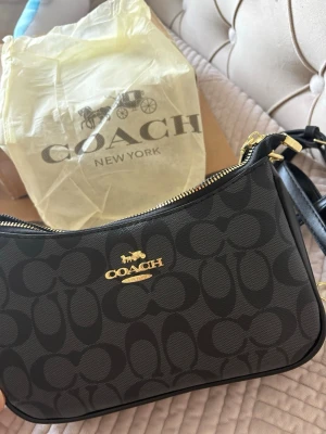 Svart axelväska från Coach - Snygg svart axelväska från Coach med klassiskt monogrammönster och guldfärgade detaljer. Väskan har justerbar axelrem och dragkedja upptill. Perfekt för dig som vill ha en stilren och trendig accessoar till din outfit.