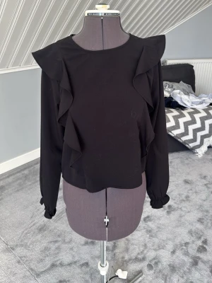 Svart blus med volanger - Säljer en elegant svart blus med volangdetaljer framtill och på axlarna. Blusen har långa ärmar med resår och liten volang vid ärmslutet. Knäpps med en knapp i nacken. Perfekt till både vardag och fest! Skick: Mycket fint skick, använd få gånger.