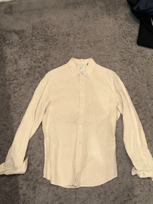 Beige linneskjorta med lång ärm - Stilren beige skjorta i linne med klassisk krage och lång ärm. Skjortan har en enkel bröstficka och knäppning framtill. Perfekt för en avslappnad men ändå snygg look.