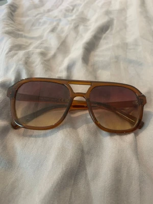Bruna retro solglasögon med brun ton - Säljer ett par snygga retroinspirerade solglasögon i brun plast med brunt tonade glas. Modellen har en markerad brygga och breda bågar som ger en cool vintagekänsla. Perfekta för dig som vill sticka ut med en unik stil.