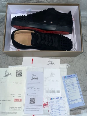 Svarta Louboutin sneakers med nitar - Svarta låga sneakers från Christian Louboutin med mattsvarta nitar över tån och sidorna. Skorna har klassisk röd sula, svarta skosnören och en stilren design i mocka. Insidan är beige och märket syns på innersulan.