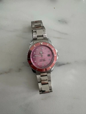 Silverfärgad klocka med rosa urtavla - Stilren klocka i silverfärgad metall med en rosa urtavla och matchande rosa ring runt glaset. Klockan har klassisk länkarmband och tydliga markeringar på urtavlan. Perfekt accessoar för dig som vill sticka ut med färg.
