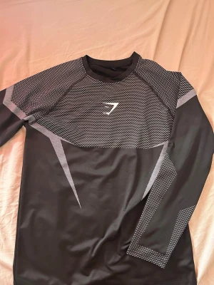 Svart långärmad träningströja Gymshark - Snygg svart långärmad träningströja från Gymshark med gråa grafiska detaljer och logga på bröstet. Tillverkad i ett stretchigt funktionsmaterial som andas, perfekt för gymmet eller löpning. Tight passform och modern design.