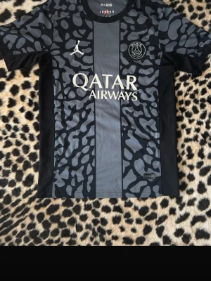 PSG x Jordan svart matchtröja - Snygg svart PSG matchtröja i samarbete med Jordan. Tröjan har ett unikt mönster i grått och svart, vita loggor och Qatar Airways tryck på bröstet. Tillverkad i lätt och ventilerande material, perfekt för fotboll eller träning.