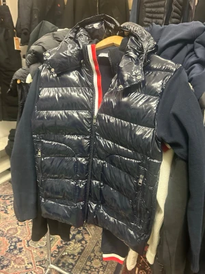 Mörkblå glansig kardigan  med huva - Snygg mörkblå moncler kardigan med glansigt material och avtagbar huva. Kardigan har dragkedja framtill, två fickor med dragkedja och quiltad design. Perfekt för lager-på-lager och streetstyle. Röd och vit randig detalj syns på insidan av kragen.