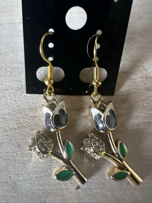 Guldiga örhängen med rosor och strass - Snygga örhängen i guldfärgad metall formade som rosor med vita och svarta detaljer. Bladen är gröna och rosorna har glittrande strass på knopparna. Perfekta för dig som vill ha något unikt och iögonfallande till din stil.
